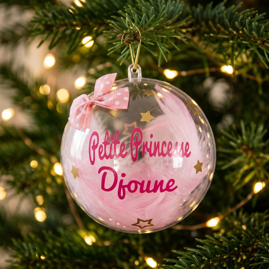 Boule de Noël transparente Petite Princesse Djoune avec plumes roses suspendue dans un sapin illuminé