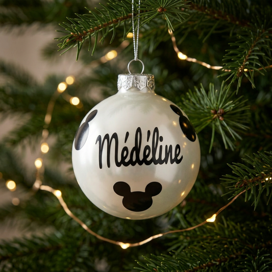 Boule de Noël blanche en verre avec prénom Médéline et motifs Mickey noirs suspendue dans un sapin illuminé